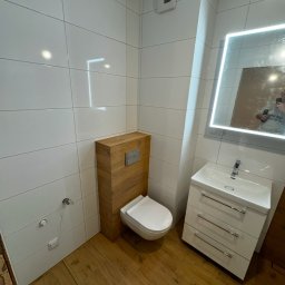 Krystian Miącz USŁUGI REMONTOWE - Nowoczesna łazienka z podwieszaną toaletą obudowaną drewnianą konstrukcją, białą umywalką z szafką i podświetlanym lustrem, wykończona białymi płytkami i drewnopodobną podłogą.