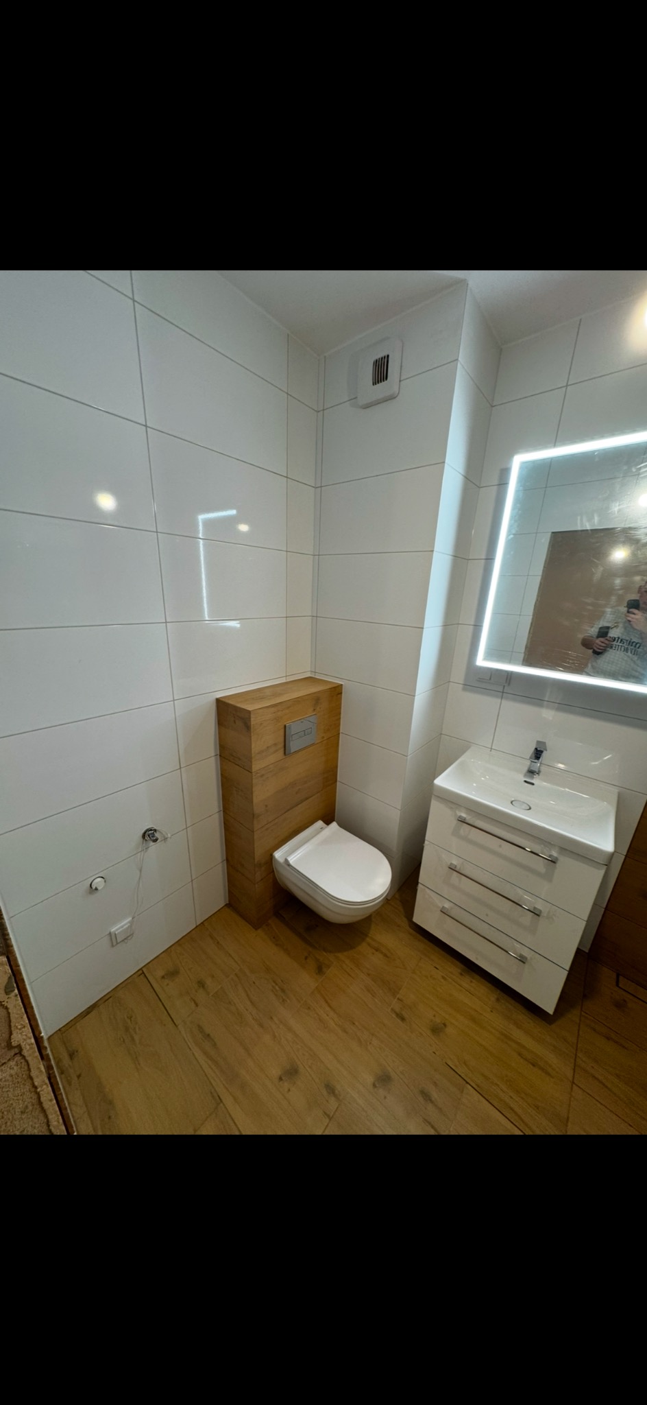 Nowoczesna łazienka z podwieszaną toaletą obudowaną drewnianą konstrukcją, białą umywalką z szafką i podświetlanym lustrem, wykończona białymi płytkami i drewnopodobną podłogą.