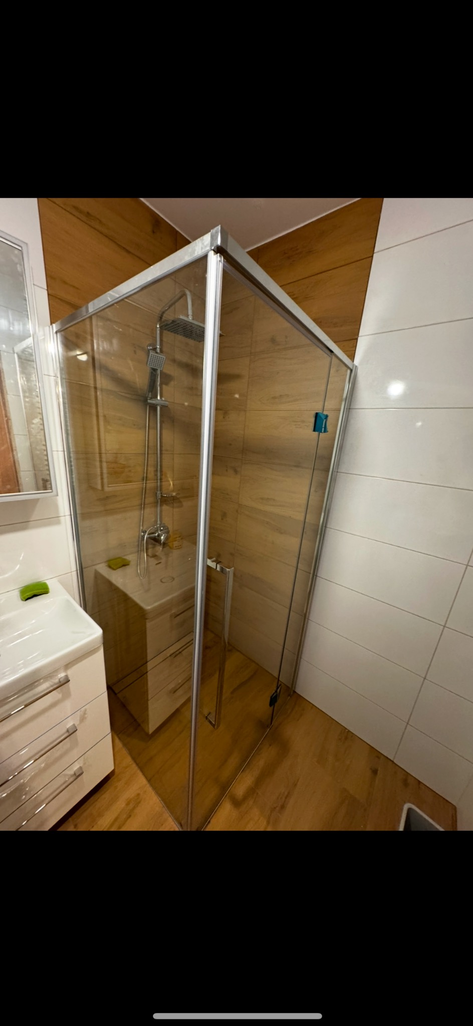 Nowoczesna kabina prysznicowa z chromowanymi profilami i deszczownicą, otoczona drewnopodobnymi i białymi płytkami, z umywalką z szafką w tle.