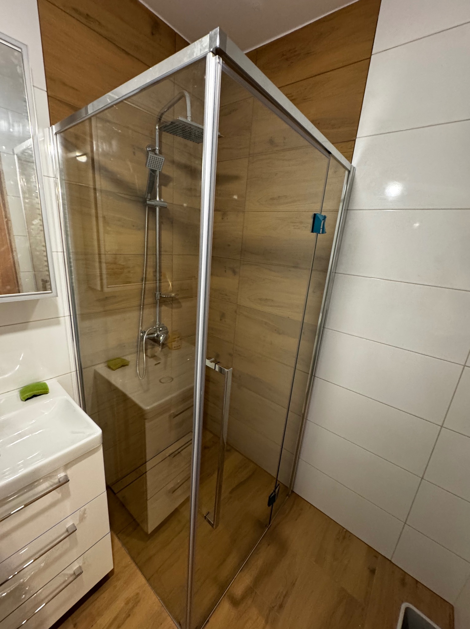 Szklana kabina prysznicowa narożna z chromowanymi profilami, widoczny brodzik z odpływem liniowym, bateria prysznicowa z deszczownicą, umywalka z szafką w tle, ściany wyłożone płytkami imitującymi...