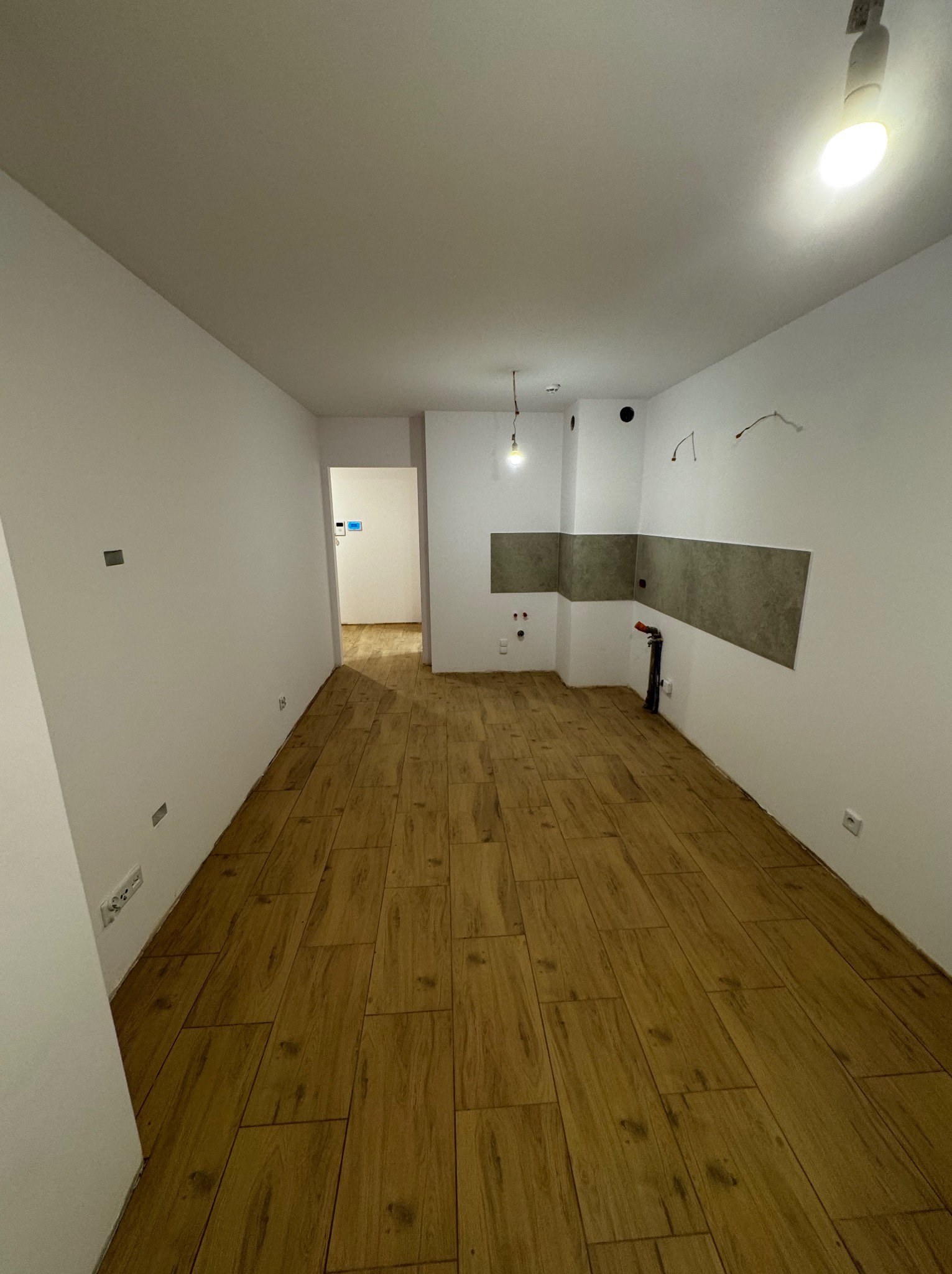 Puste pomieszczenie po remoncie z drewnopodobną podłogą, świeżo pomalowanymi białymi ścianami i przygotowanym miejscem na kuchnię z szarymi płytkami nad blatem oraz wystającymi instalacjami.