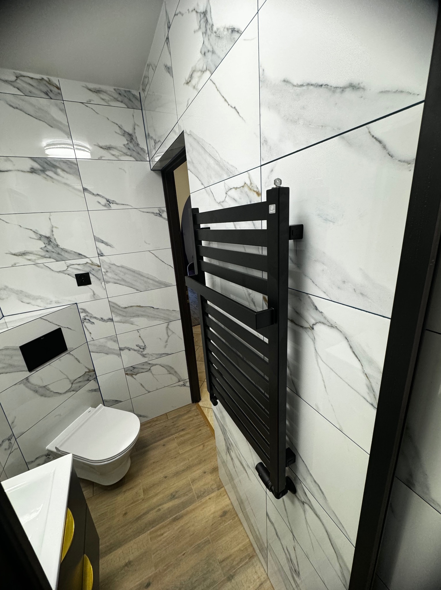 Nowoczesna łazienka z białymi płytkami imitującymi marmur, drewnopodobną podłogą i czarnym grzejnikiem drabinkowym oraz podwieszaną toaletą.
