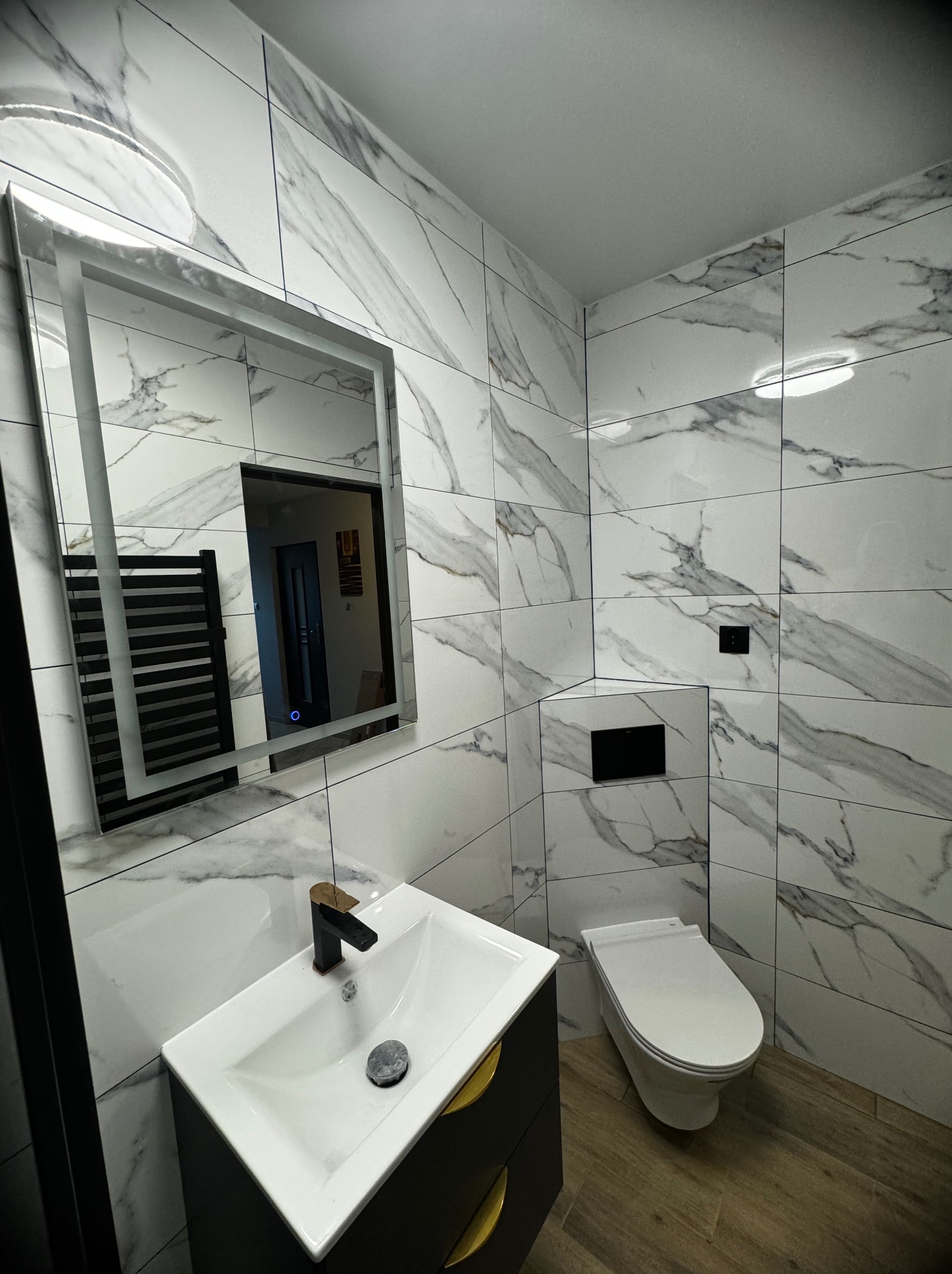Nowoczesna łazienka z białymi płytkami imitującymi marmur, podwieszana toaleta, umywalka nablatowa z czarną armaturą i szafką z złotymi uchwytami, podświetlane lustro.