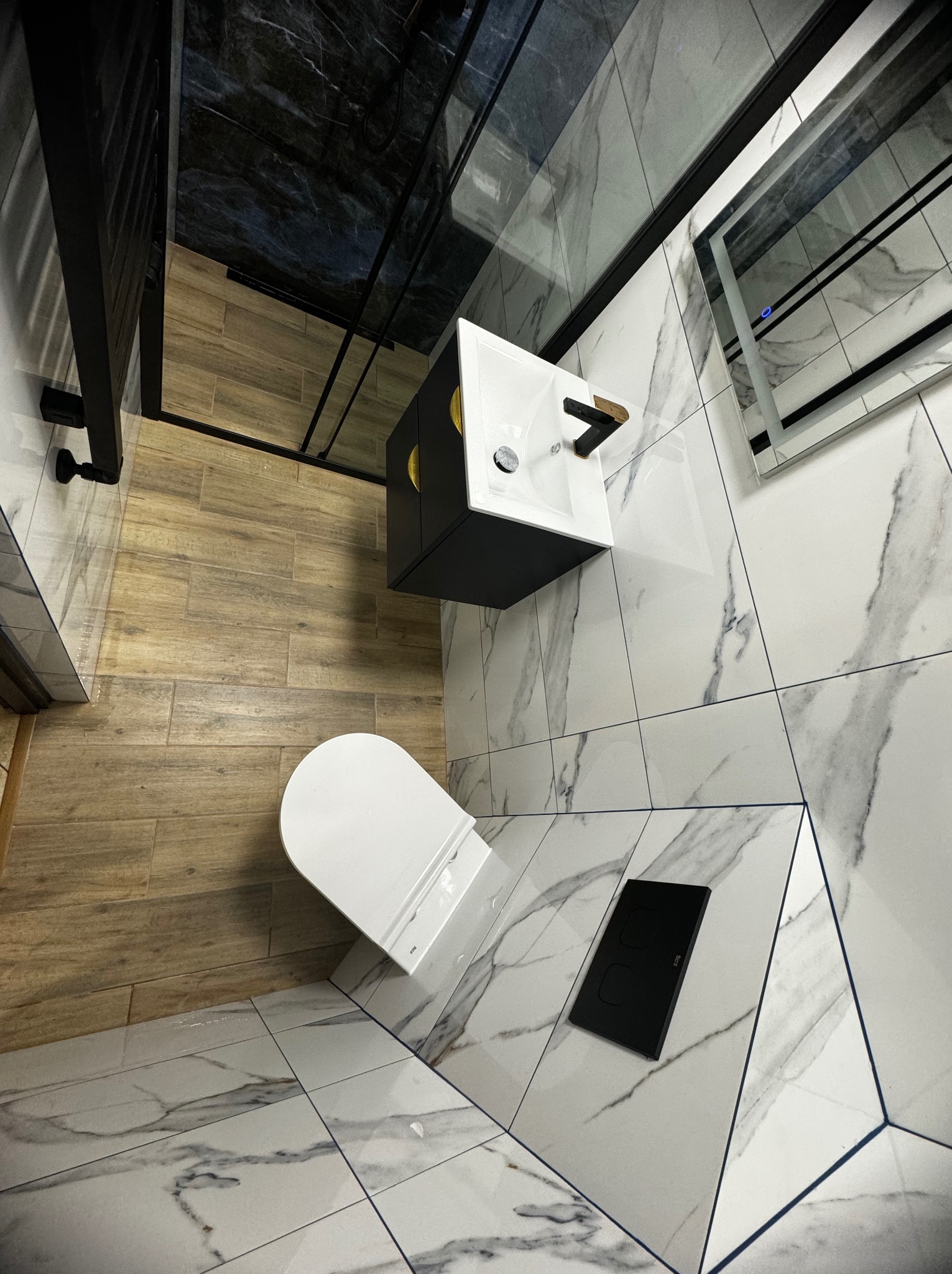 Nowoczesna łazienka po remoncie z marmurowymi płytkami na ścianach i podłodze, drewnopodobnymi płytkami w strefie prysznica, czarną armaturą i podwieszaną szafką umywalkową z złotymi akcentami.