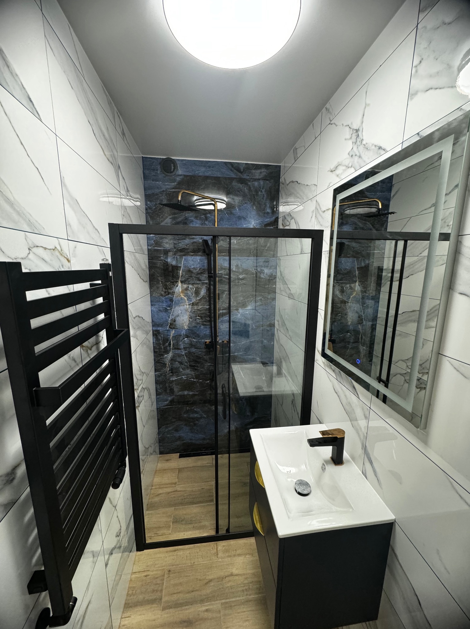 Nowoczesna łazienka z kabiną prysznicową typu walk-in, umywalką nablatową z czarną armaturą i podświetlanym lustrem, wykończona białymi płytkami imitującymi marmur.