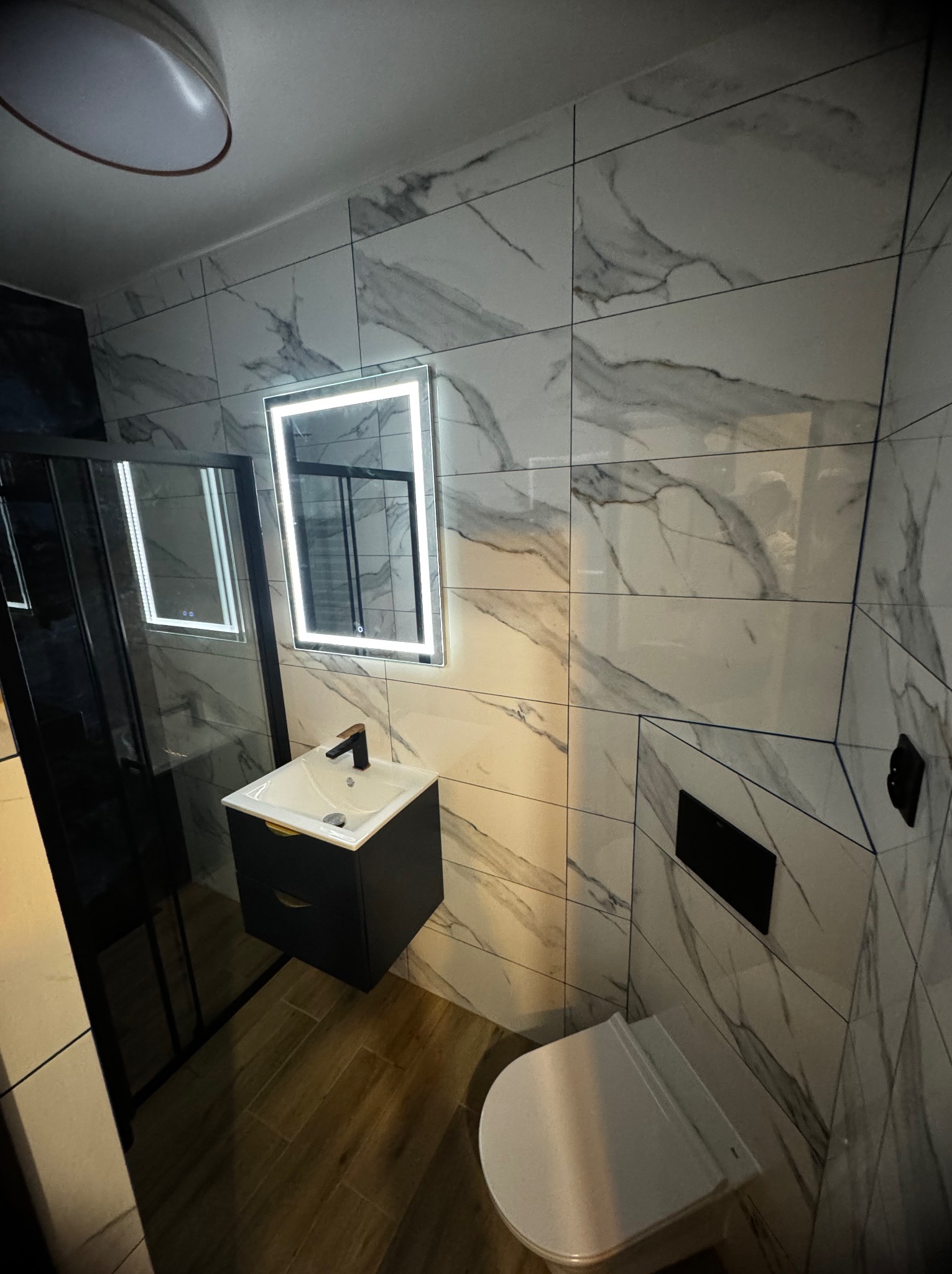 Nowoczesna łazienka z marmurowymi płytkami, podświetlanym lustrem, czarną szafką pod umywalką i kabiną prysznicową z czarnymi ramami.