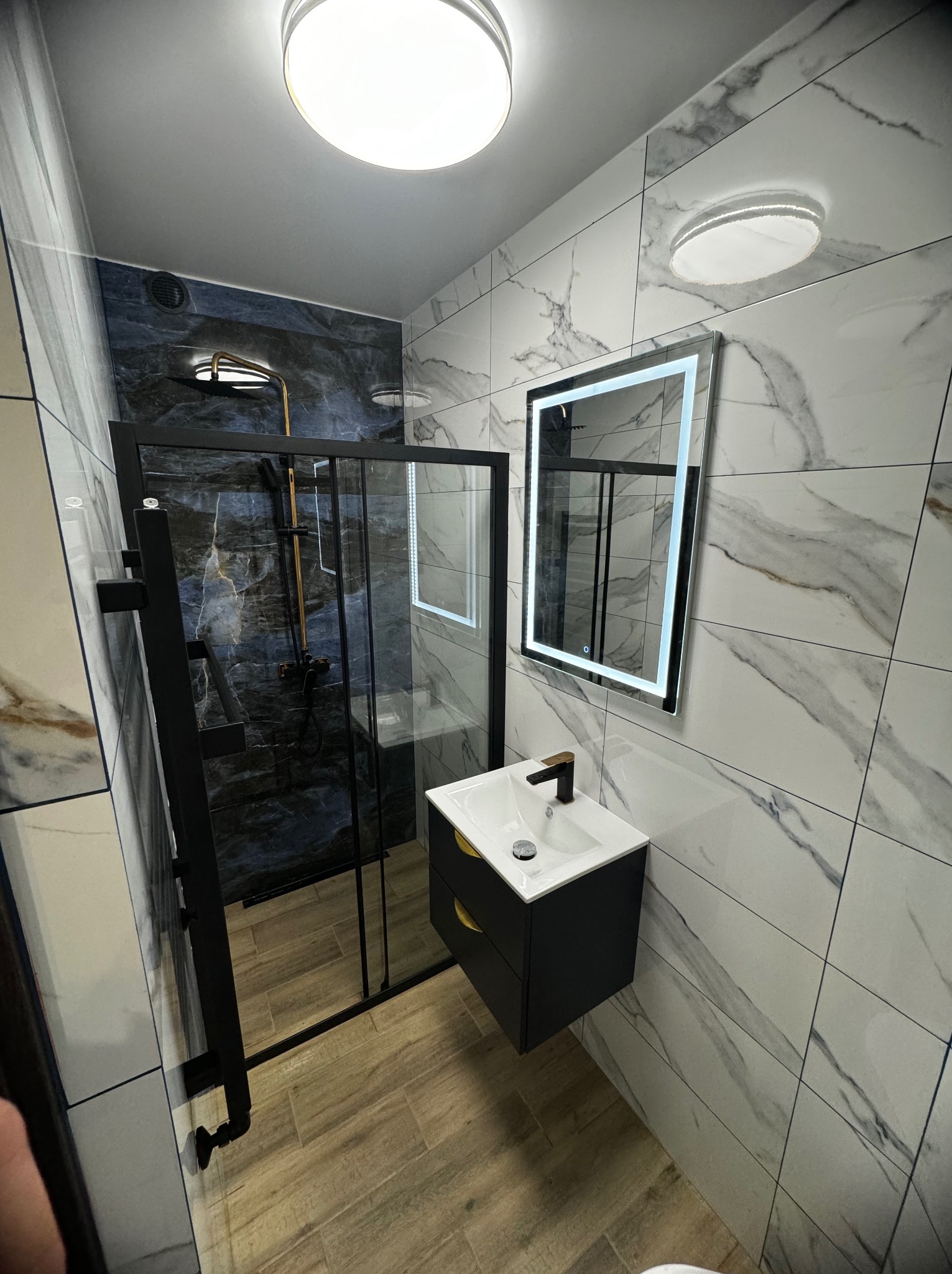 Nowoczesna łazienka z czarną kabiną prysznicową, umywalką z szafką w kolorze antracytu i podświetlanym lustrem na ścianie wyłożonej płytkami imitującymi marmur.