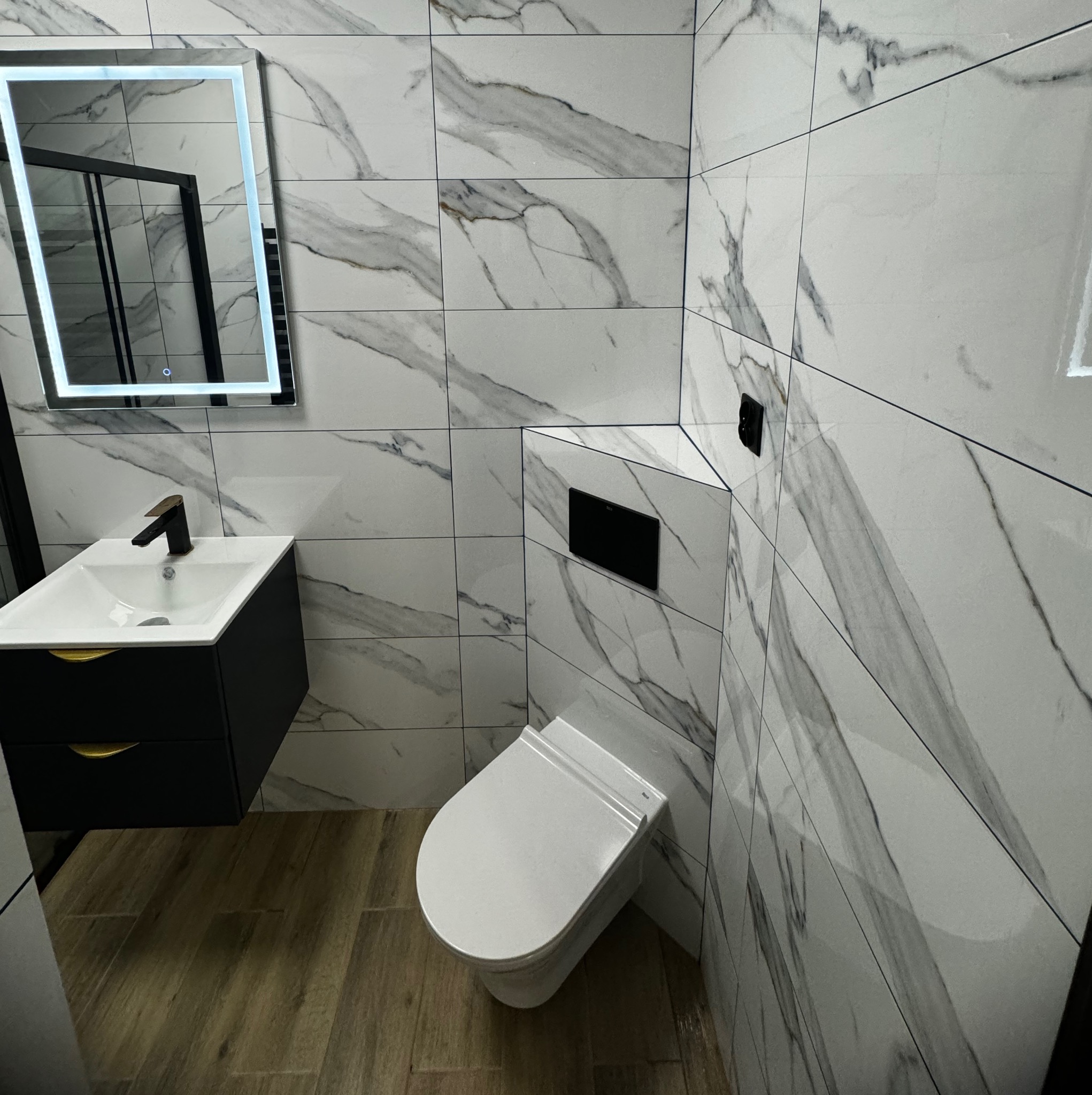 Nowoczesna łazienka z białymi płytkami imitującymi marmur, podwieszana toaleta, szafka z umywalką w kolorze czarnym z złotymi uchwytami i podświetlane lustro.