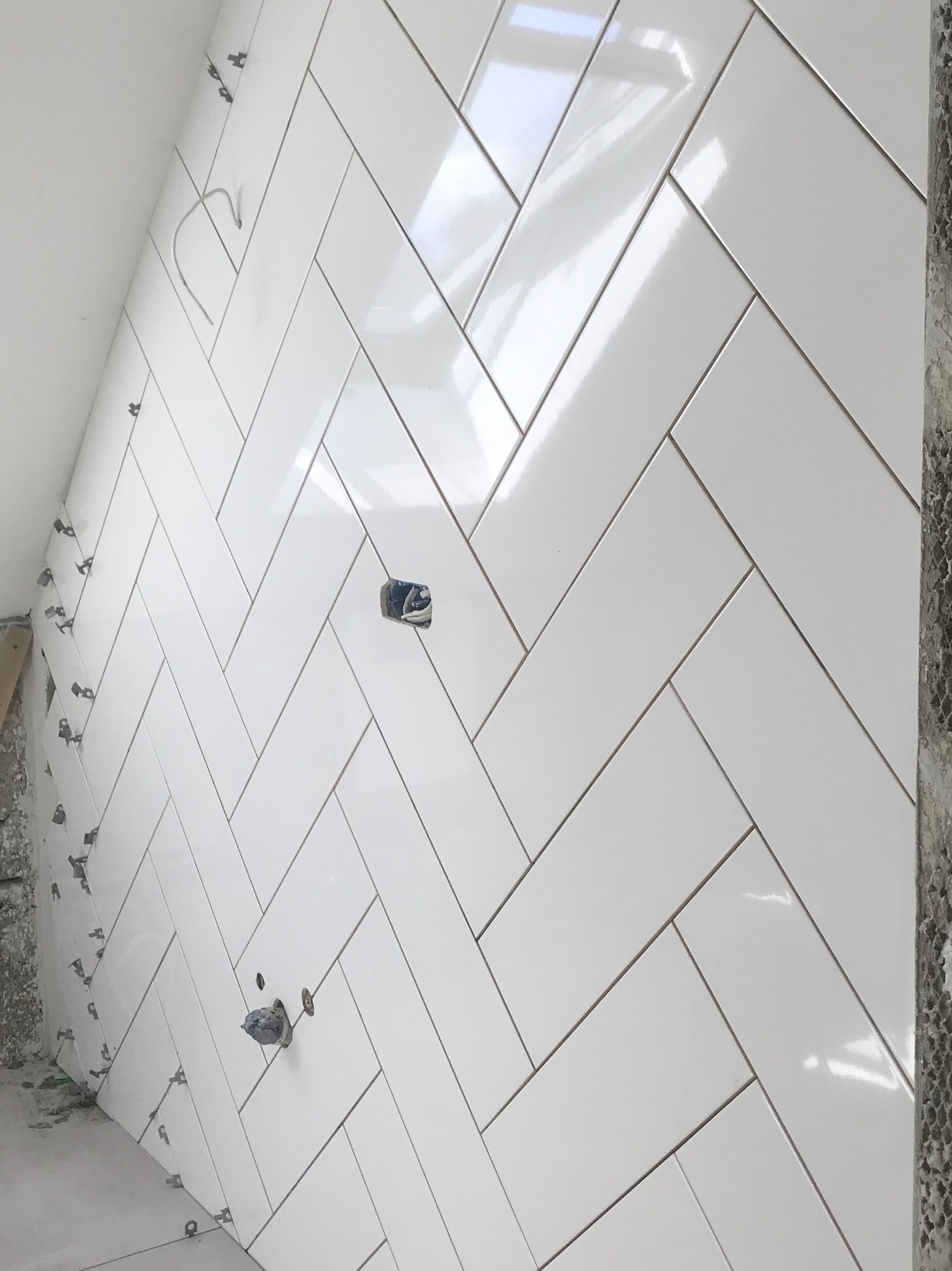 Białe, błyszczące płytki ułożone w jodełkę na ścianie, z widocznymi krzyżykami dystansowymi i otworami na gniazdka elektryczne w trakcie montażu.