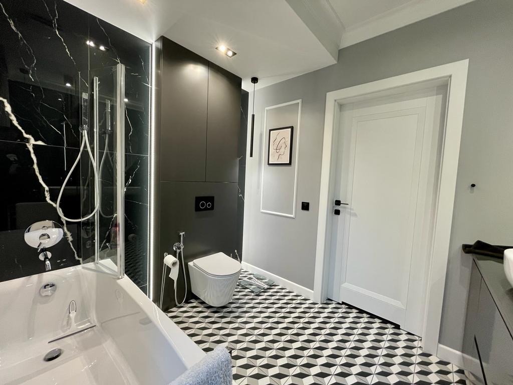Nowoczesna łazienka z czarno-białą mozaiką na podłodze, czarnymi płytkami imitującymi marmur na ścianie przy wannie, wiszącą toaletą i minimalistyczną grafiką na szarej ścianie.