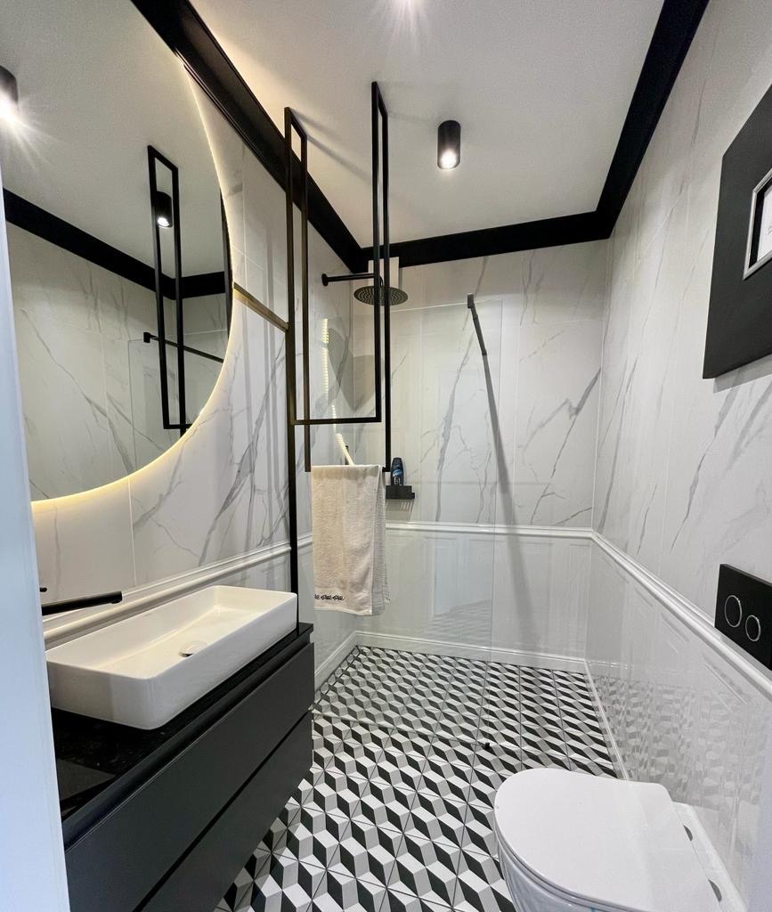 Nowoczesna łazienka z biało-czarną mozaiką podłogową, marmurowymi płytkami ściennymi, podświetlanym lustrem owalnym, czarną armaturą i kabiną prysznicową ze szklaną ścianką.