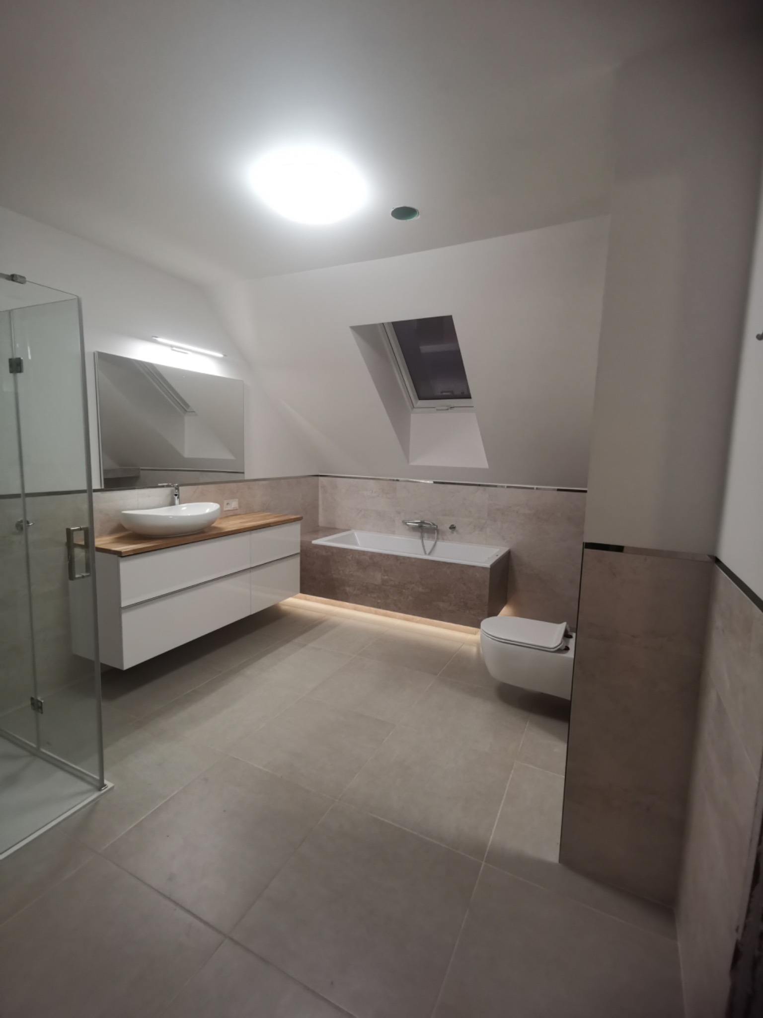 Nowoczesna łazienka na poddaszu z oknem dachowym, beżowe płytki, podświetlana wanna, biała umywalka nablatowa na drewnianym blacie i szklana kabina prysznicowa.