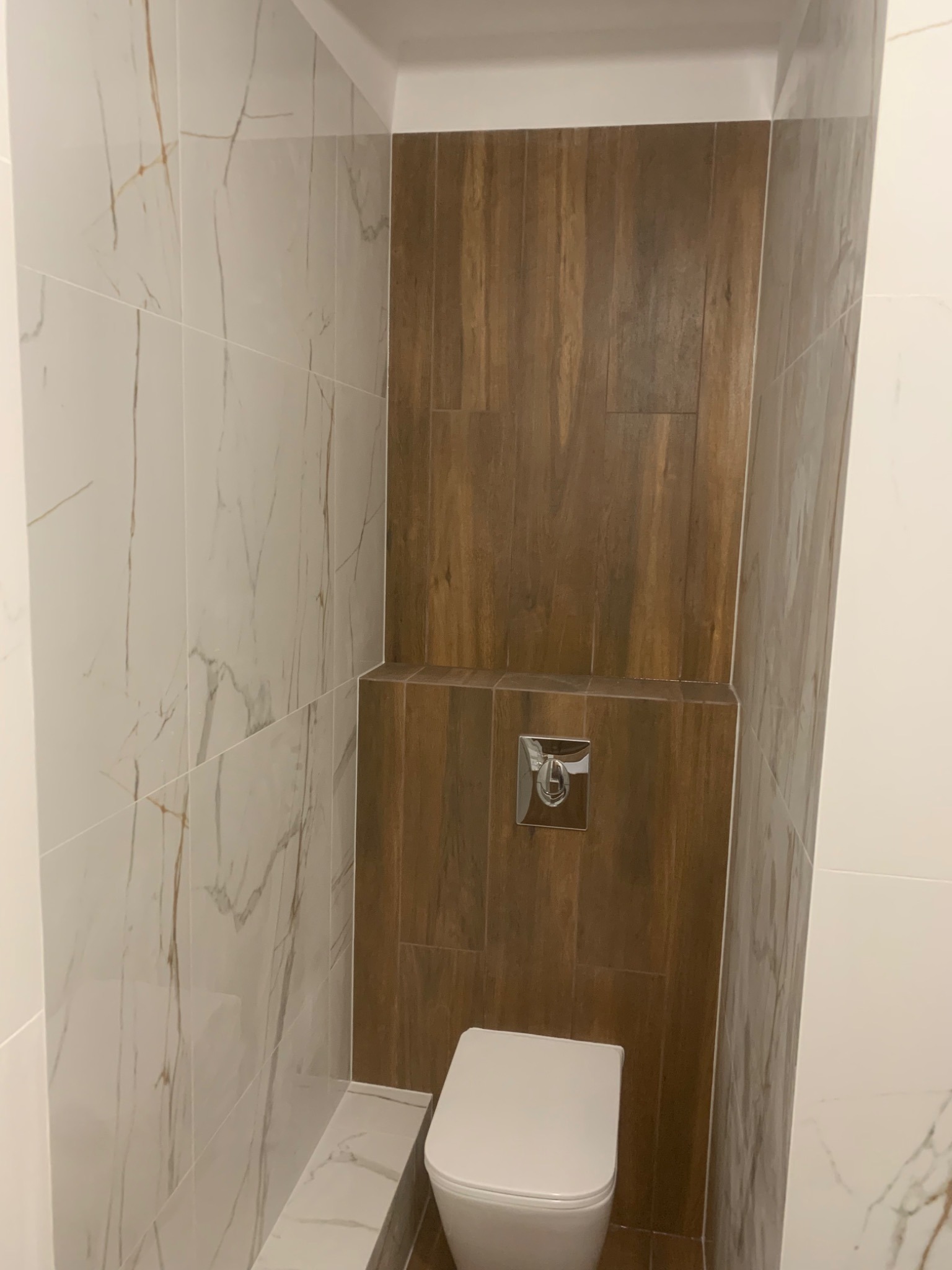 Nowoczesna toaleta podwieszana w zabudowie z płytek imitujących drewno, kontrastująca z marmurowymi płytkami na ścianach, chromowany przycisk spłukiwania.