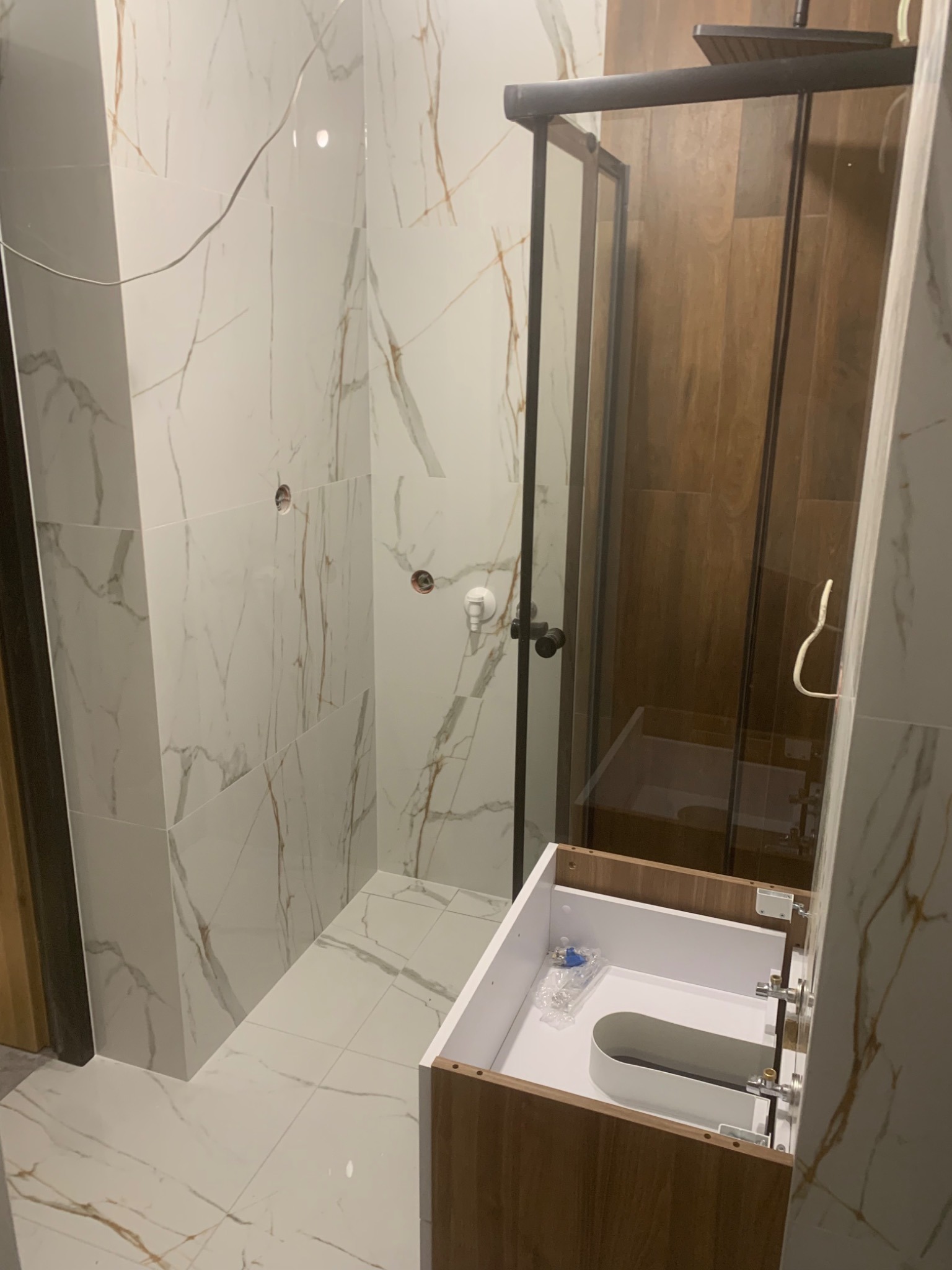 Nowoczesna łazienka w trakcie wykańczania: kabina prysznicowa z czarnymi ramami, białe płytki imitujące marmur z brązowymi żyłami, drewnopodobna szafka pod umywalkę w stanie surowym, widoczne...