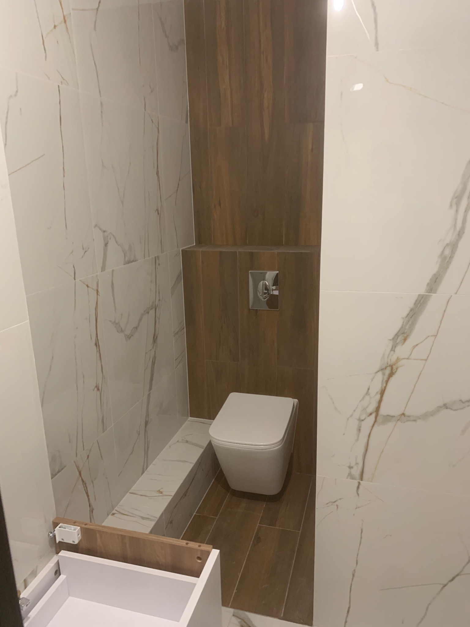 Nowoczesna łazienka w trakcie wykańczania: białe płytki imitujące marmur, drewnopodobne płytki, wisząca toaleta z chromowanym przyciskiem spłukiwania oraz niedokończona szafka pod umywalkę.