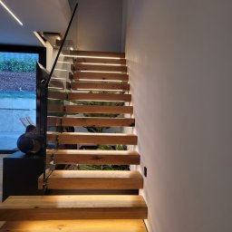 Usługi Elektryczne Damian Zygmunt - Nowoczesne schody drewniane z podświetleniem LED wbudowanym w stopnie, szklana balustrada, widok z dołu, obok minimalistyczny głośnik.