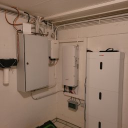 Usługi Elektryczne Damian Zygmunt - Szara skrzynka elektryczna z drzwiczkami, obok białe urządzenia SolarEdge na ścianie w piwnicy, widoczne kable i rurki instalacyjne pod sufitem, narzędzia w zielonej skrzynce na podłodze.