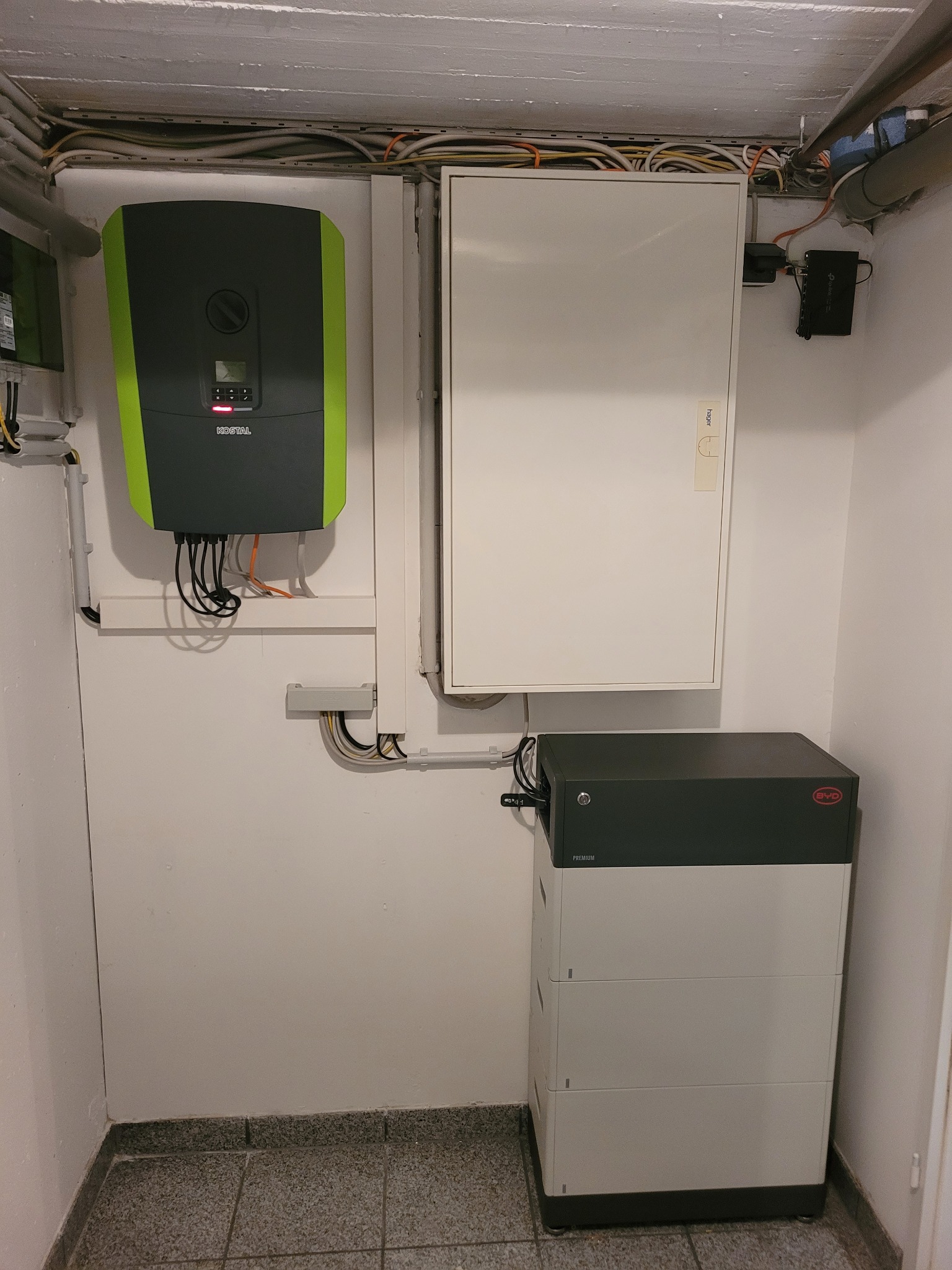 Zestaw instalacji fotowoltaicznej w piwnicy: inwerter Kostal z zieloną obudową, akumulator BYD, skrzynka elektryczna Hager i kable prowadzone pod sufitem.