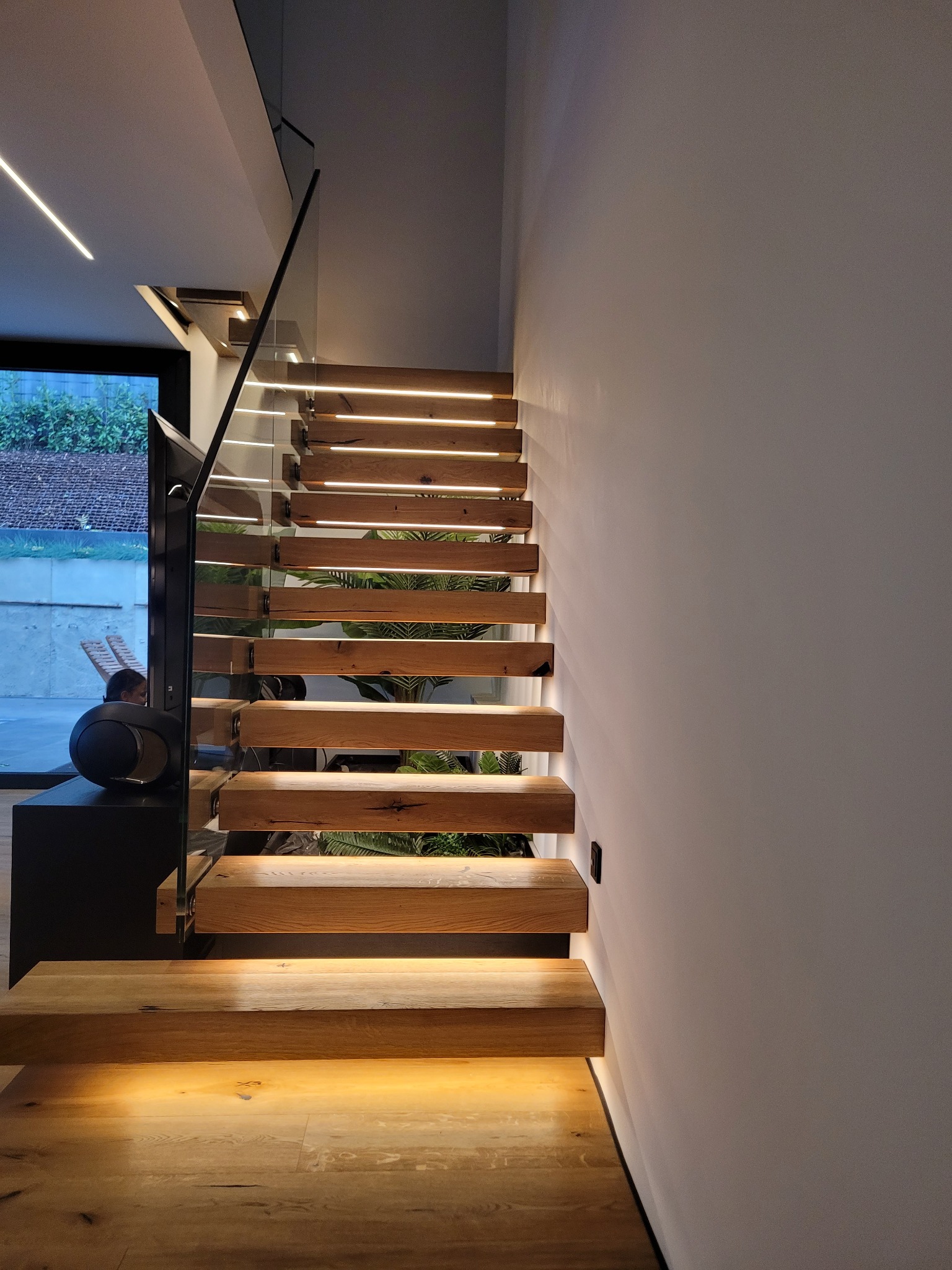 Nowoczesne schody drewniane z podświetleniem LED wbudowanym w stopnie, szklana balustrada, widok z dołu, obok minimalistyczny głośnik.