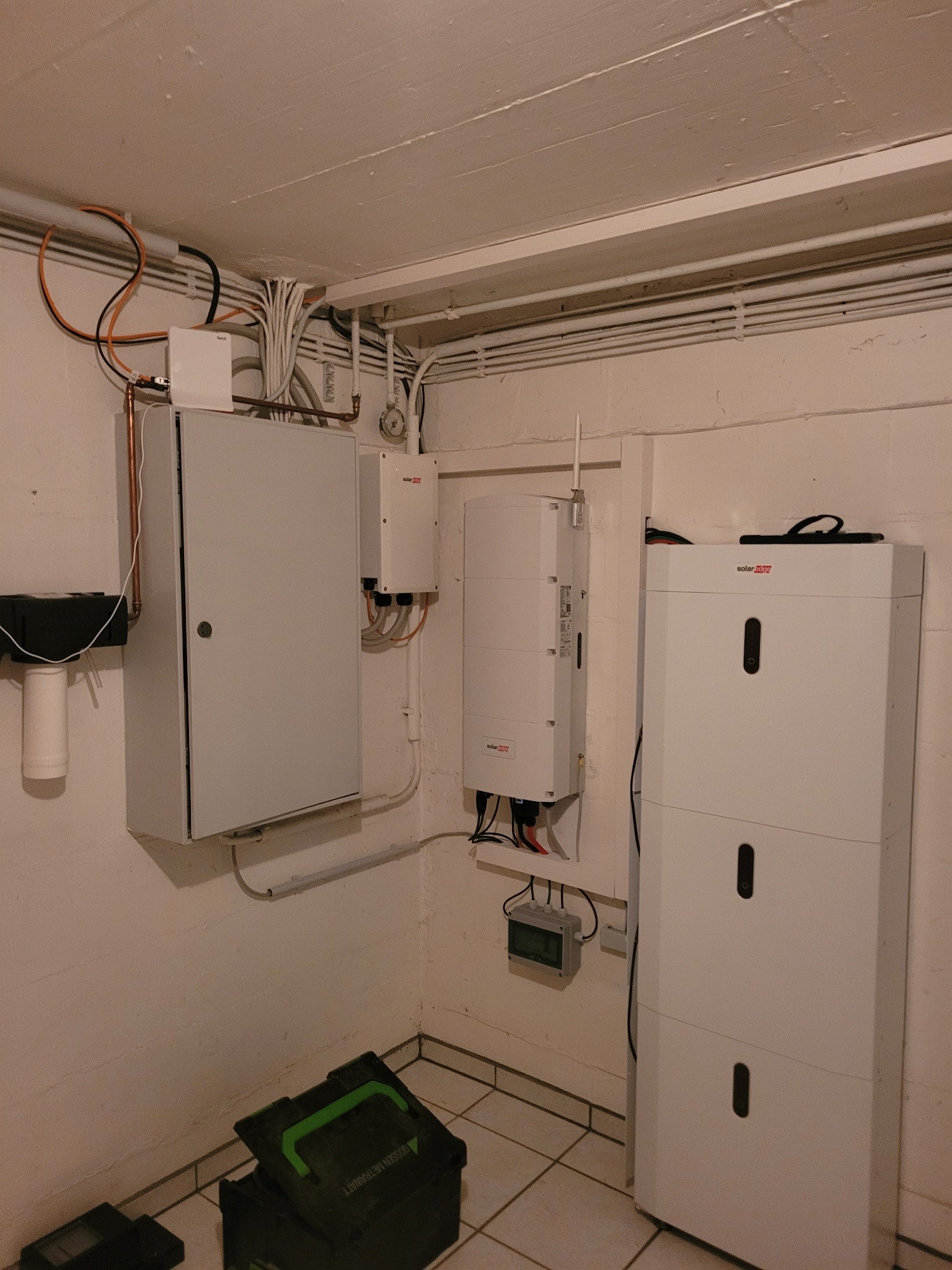 Szara skrzynka elektryczna z drzwiczkami, obok białe urządzenia SolarEdge na ścianie w piwnicy, widoczne kable i rurki instalacyjne pod sufitem, narzędzia w zielonej skrzynce na podłodze.