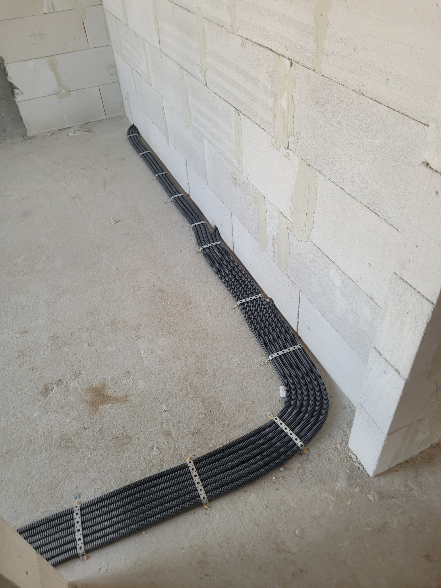 Kable elektryczne w peszlach ułożone wzdłuż ściany z betonu komórkowego, przymocowane metalowymi uchwytami z otworami, surowy stan wykończenia wnętrza.