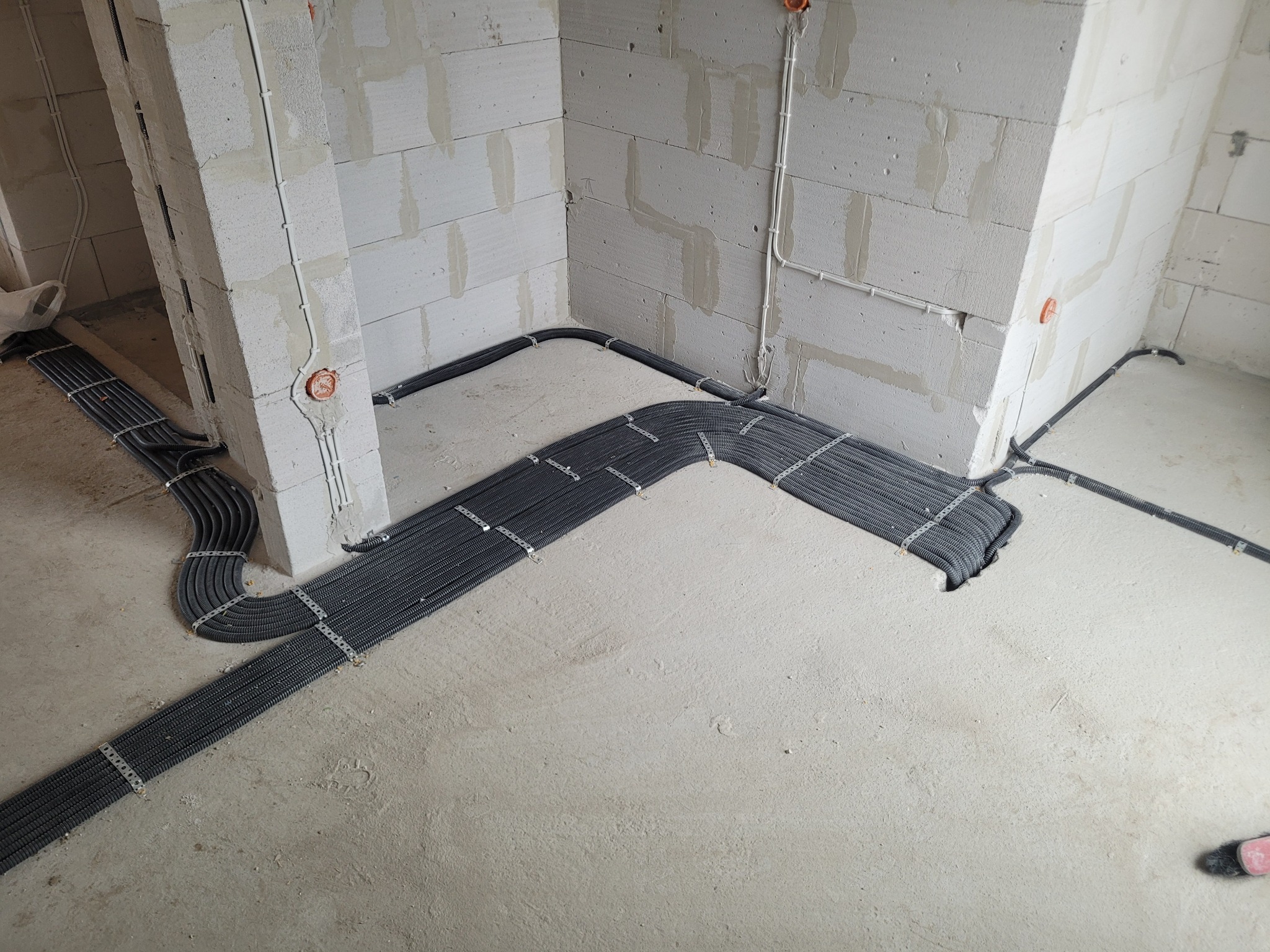 Szare, karbowane rury elektryczne ułożone równolegle i przymocowane metalowymi uchwytami do surowego, betonowego podłoża w trakcie instalacji w nowym budynku z białych bloczków.