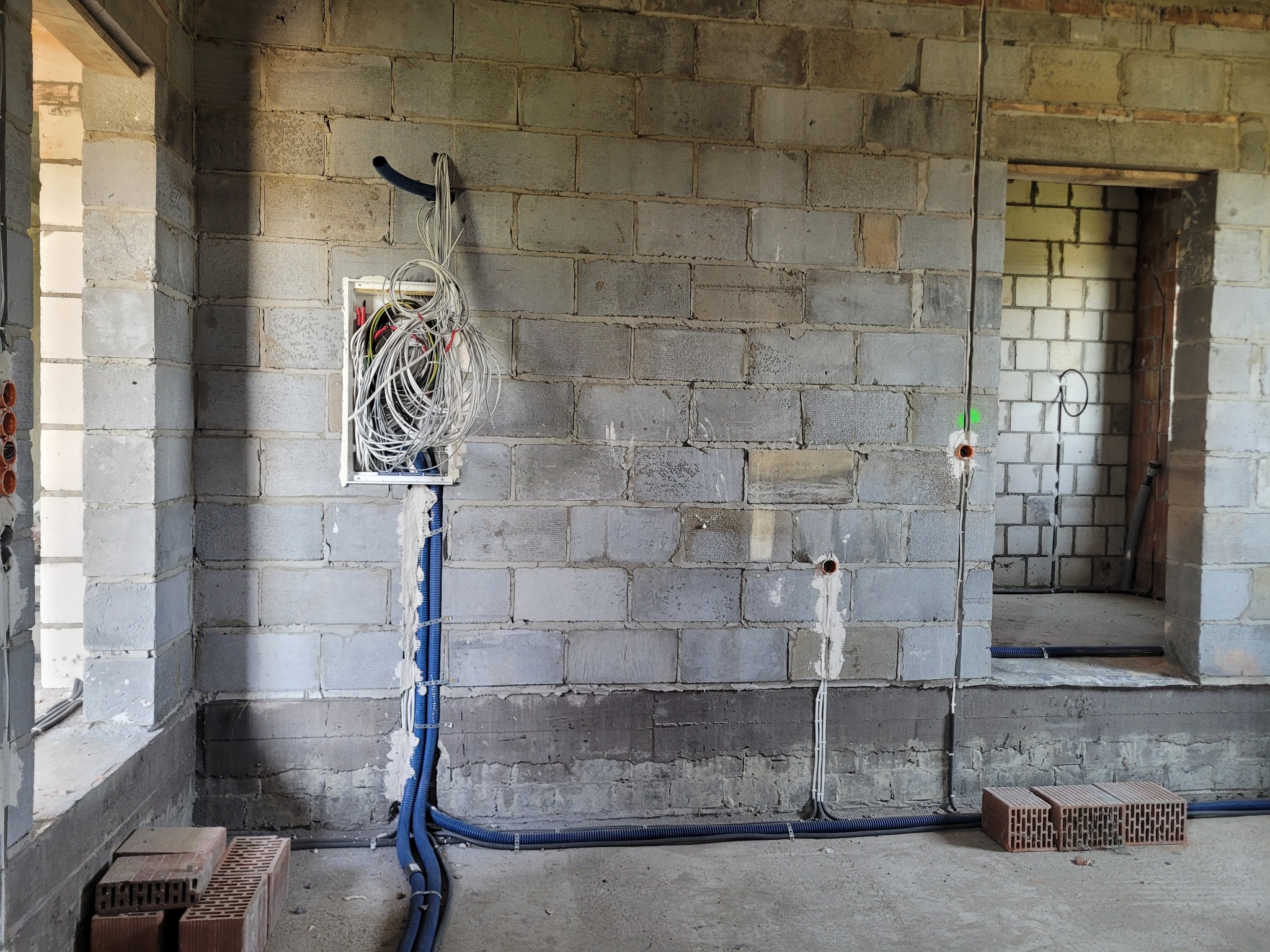 Surowa ściana z betonu komórkowego w trakcie prac wykończeniowych, widoczna puszka podtynkowa z nieuporządkowanymi przewodami elektrycznymi, niebieskie rury karbowane prowadzące w dół ściany...