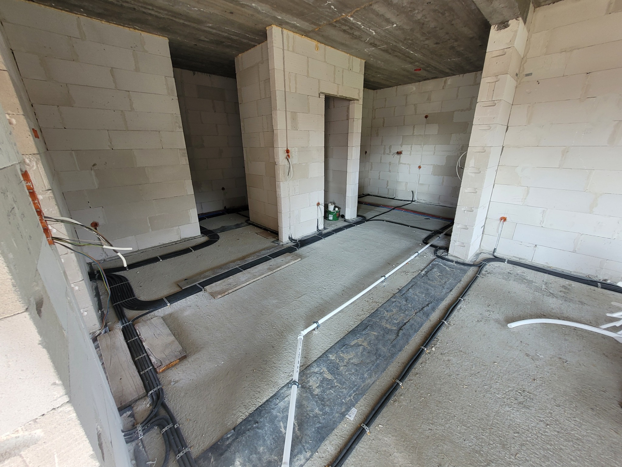 Surowy stan wnętrza z instalacją elektryczną w peszlach ułożoną na podłodze, widoczne przewody wychodzące ze ściany i puszki instalacyjne.