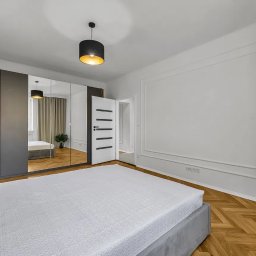 Angelika Bujakowska - Nowoczesna sypialnia z parkietem w jodełkę, szarą szafą z lustrem i minimalistyczną lampą wiszącą. W tle widoczne otwarte drzwi.