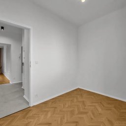 Angelika Bujakowska - Jasne wnętrze: parkiet w jodełkę, białe ściany, roślina w koszu i widok na korytarz z beżowymi zasłonami. Minimalistyczny design, świeżo po remoncie.