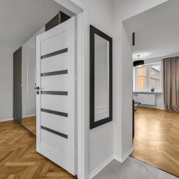 Angelika Bujakowska - Jasne wnętrze mieszkania z parkietem w jodełkę, białe drzwi z czarnymi paskami i lustro w czarnej ramie. W tle widoczne okno z zasłonami i roślina doniczkowa.