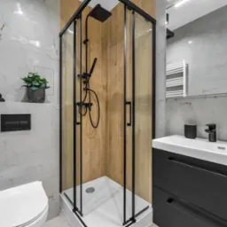 Nowoczesna łazienka z kabiną prysznicową w czarnej ramie, umywalką z czarną szafką i armaturą. Marmurowe płytki, drewniane akcenty i roślina doniczkowa.