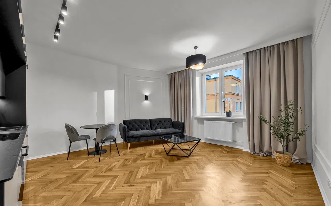 Jasne wnętrze z parkietem w jodełkę, szarą sofą i stołem. Okno z widokiem na budynek. Nowoczesne oświetlenie sufitowe i dekoracyjne detale.