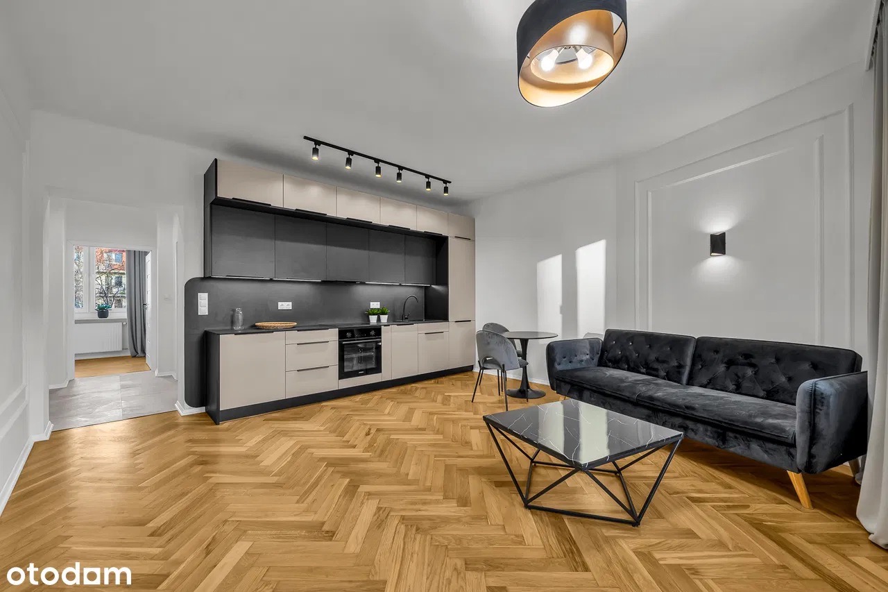 Nowoczesne wnętrze: parkiet w jodełkę, czarno-beżowa kuchnia z oświetleniem szynowym, ciemna sofa i stolik kawowy. W głębi widoczne kolejne pomieszczenie.