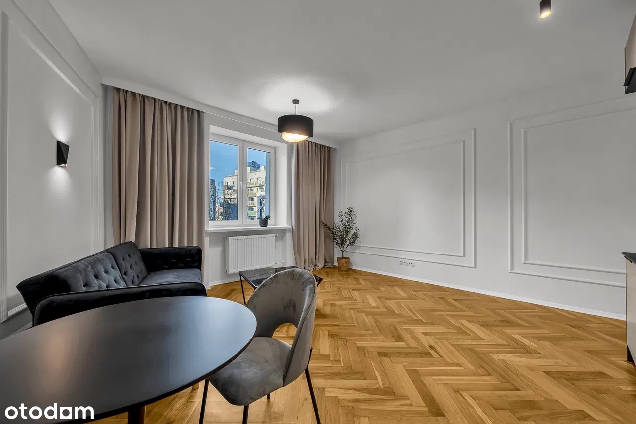 Jasne wnętrze: parkiet w jodełkę, czarna sofa i stół, szare krzesło. Okno z widokiem na miasto. Dekoracyjne listwy na ścianach i minimalistyczne oświetlenie.