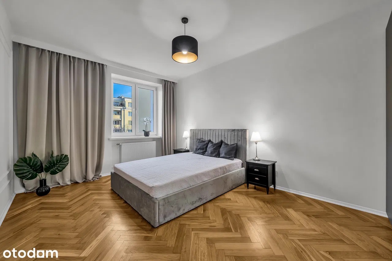 Sypialnia w jasnych barwach z parkietem w jodełkę. Szare zasłony, łóżko z welurowym zagłówkiem, czarne szafki nocne i lampki. Za oknem widok na budynek.