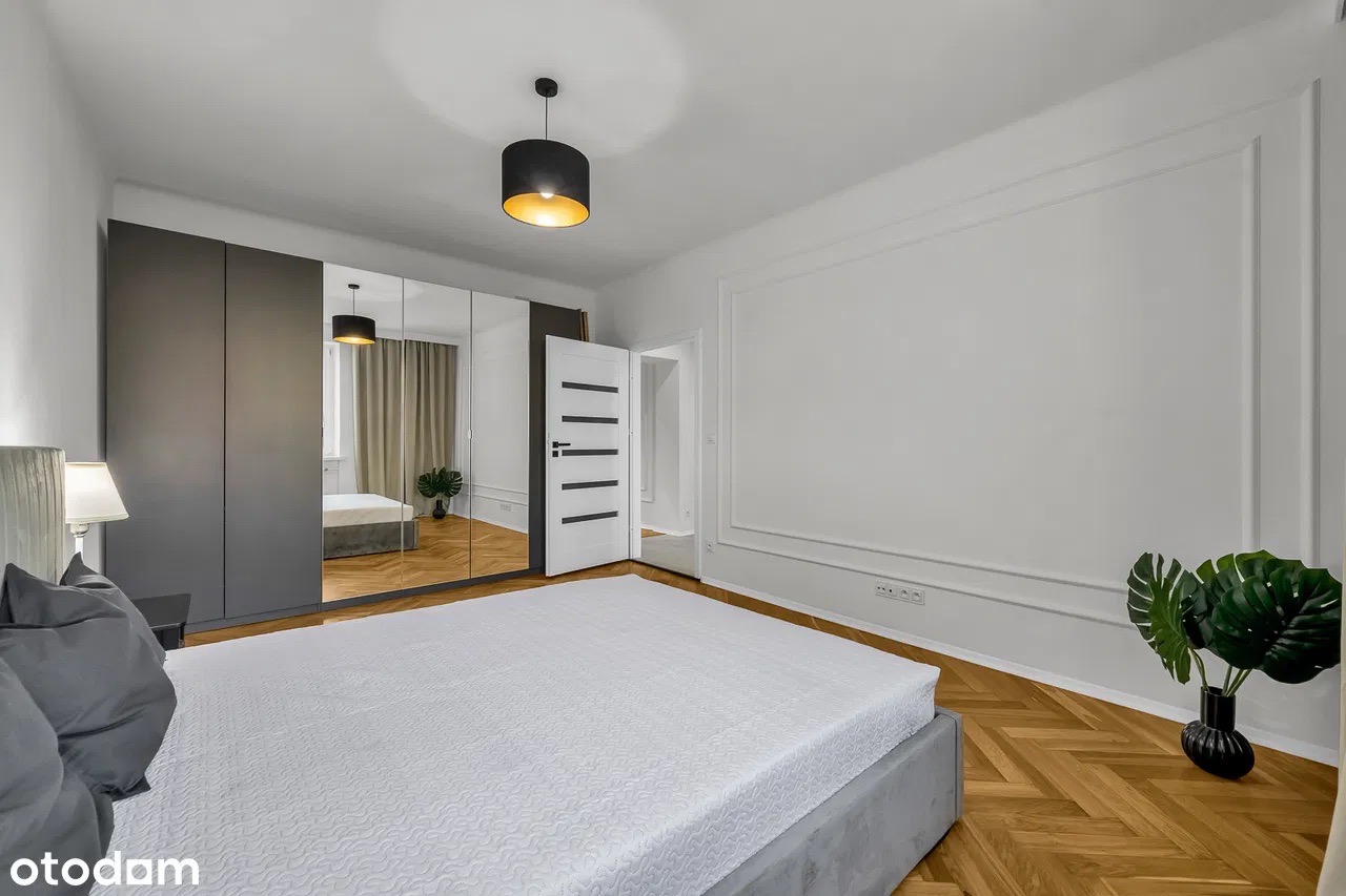 Nowoczesna sypialnia z parkietem w jodełkę, szarą szafą z lustrem i minimalistyczną lampą wiszącą. W tle widoczne otwarte drzwi.