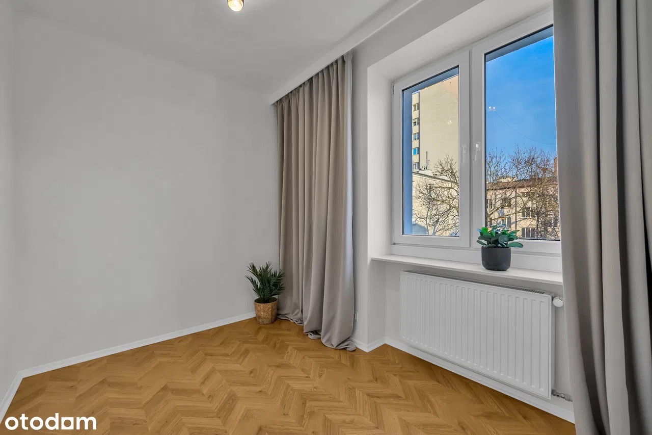 Jasne wnętrze z parkietem w jodełkę, grzejnikiem i oknem z widokiem na budynki. Szare zasłony i roślina doniczkowa dodają przytulności. Minimalistyczny design.