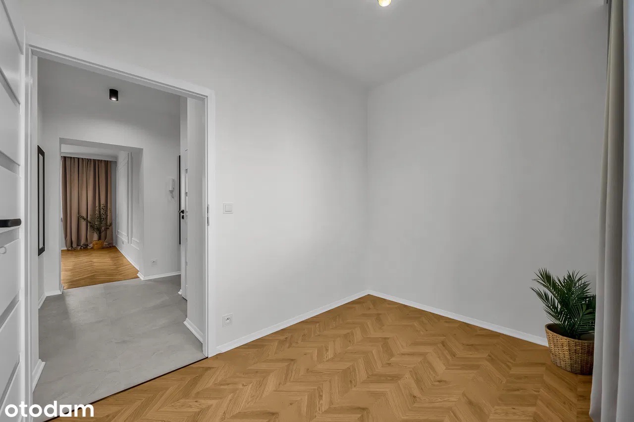 Jasne wnętrze: parkiet w jodełkę, białe ściany, roślina w koszu i widok na korytarz z beżowymi zasłonami. Minimalistyczny design, świeżo po remoncie.