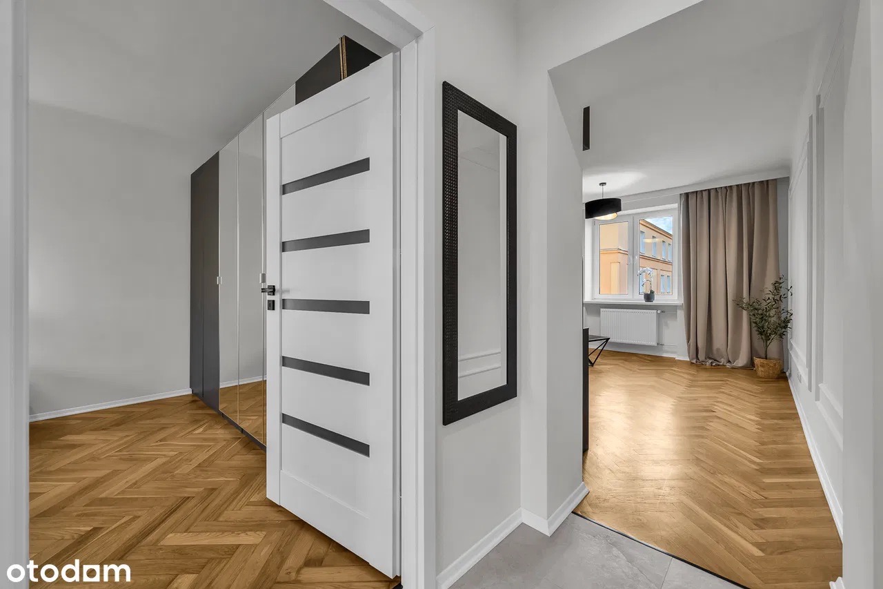 Jasne wnętrze mieszkania z parkietem w jodełkę, białe drzwi z czarnymi paskami i lustro w czarnej ramie. W tle widoczne okno z zasłonami i roślina doniczkowa.