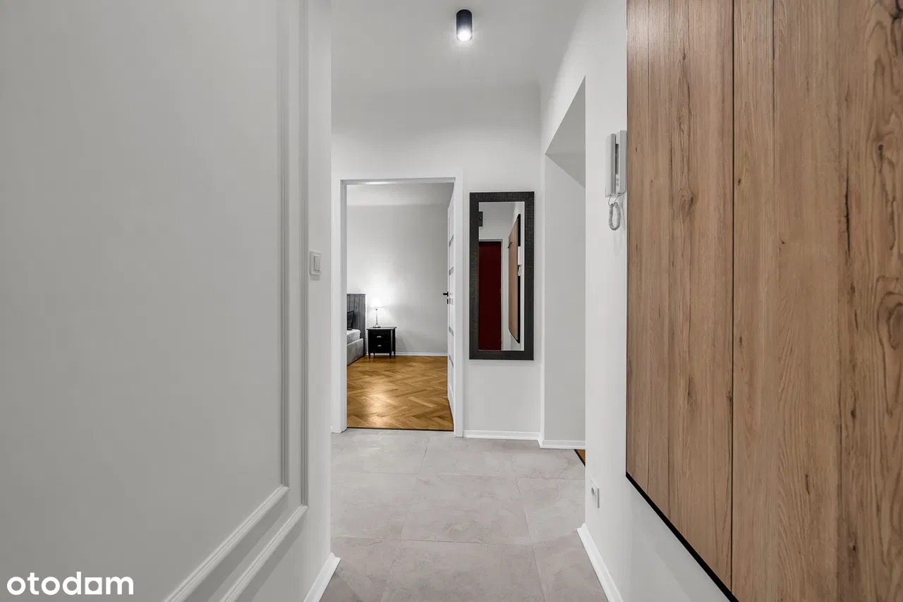 Nowoczesny korytarz z jasnymi ścianami, szarą podłogą i drewnianą szafą. W głębi widok na sypialnię z parkietem i czarną szafką nocną.