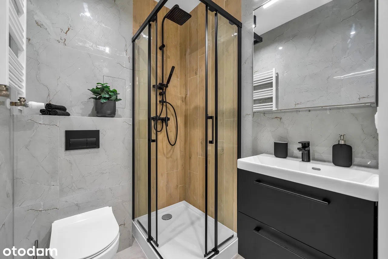 Nowoczesna łazienka z kabiną prysznicową w czarnej ramie, umywalką z czarną szafką i armaturą. Marmurowe płytki, drewniane akcenty i roślina doniczkowa.