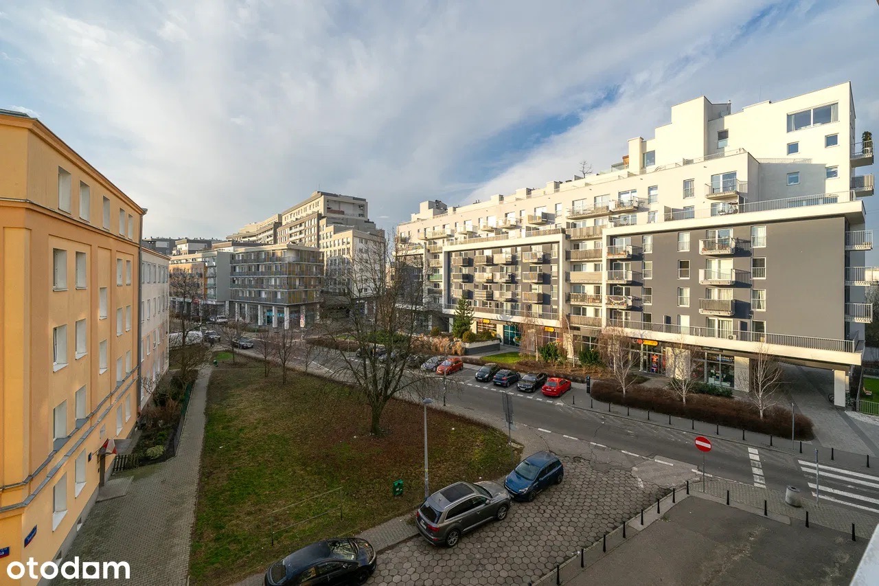 Widok z balkonu na nowoczesne osiedle z budynkami o zróżnicowanej architekturze, zielenią i zaparkowanymi samochodami w słoneczny dzień.