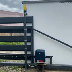 Zamontowany napęd do bramy automatycznej BFT Ultra z lampą ostrzegawczą na słupku, widoczne przewody i podłączenie, tło stanowi biała ściana budynku i fragment dachu innego budynku.