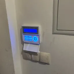 Panel alarmowy z podświetlanym wyświetlaczem LCD i klawiaturą, zamontowany na ścianie obok włączników światła i skrzynki elektrycznej.
