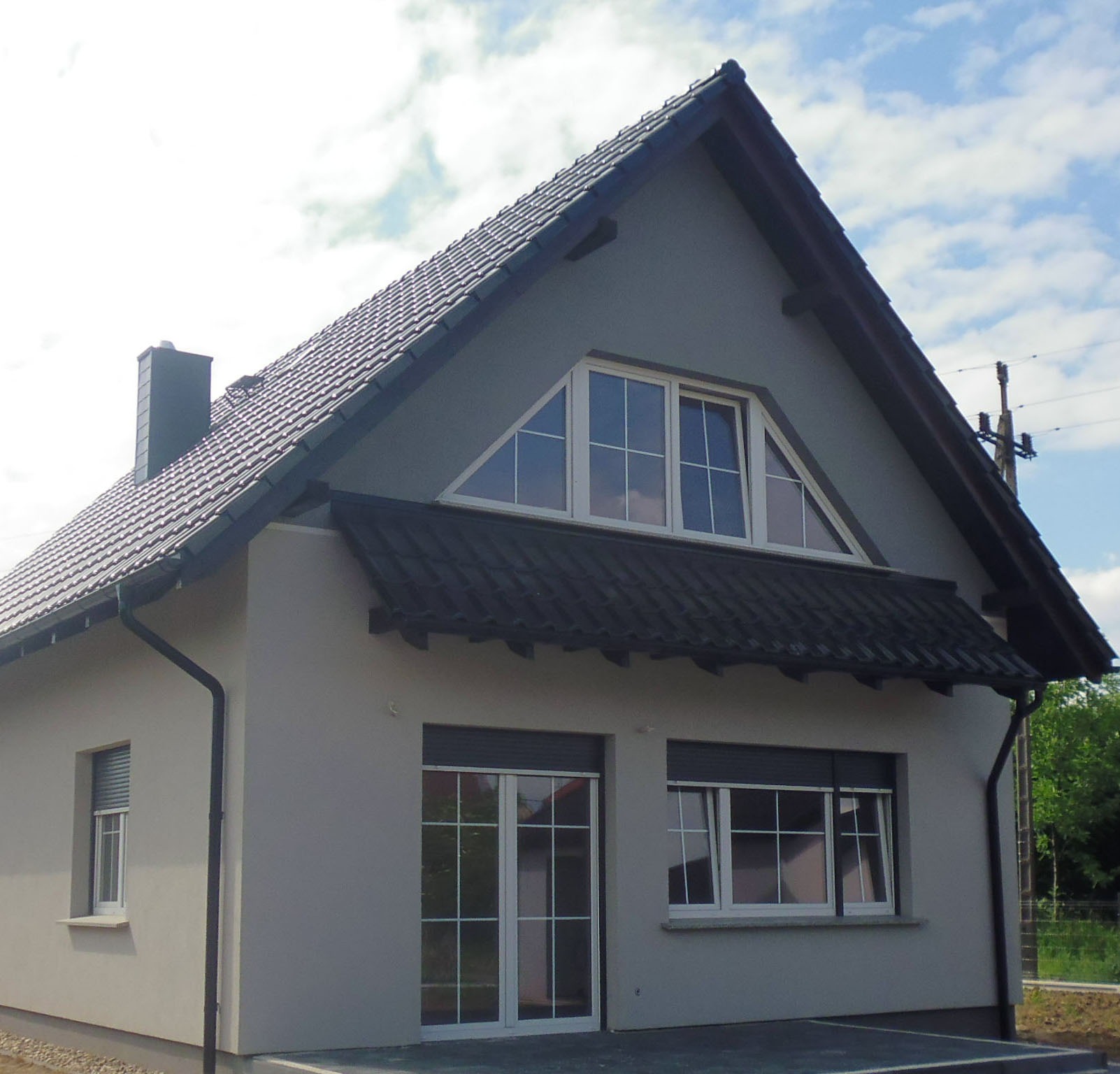 Narożny widok parterowego domu z szarą elewacją, ciemną dachówką, białymi oknami z roletami i trójkątnym oknem na poddaszu.