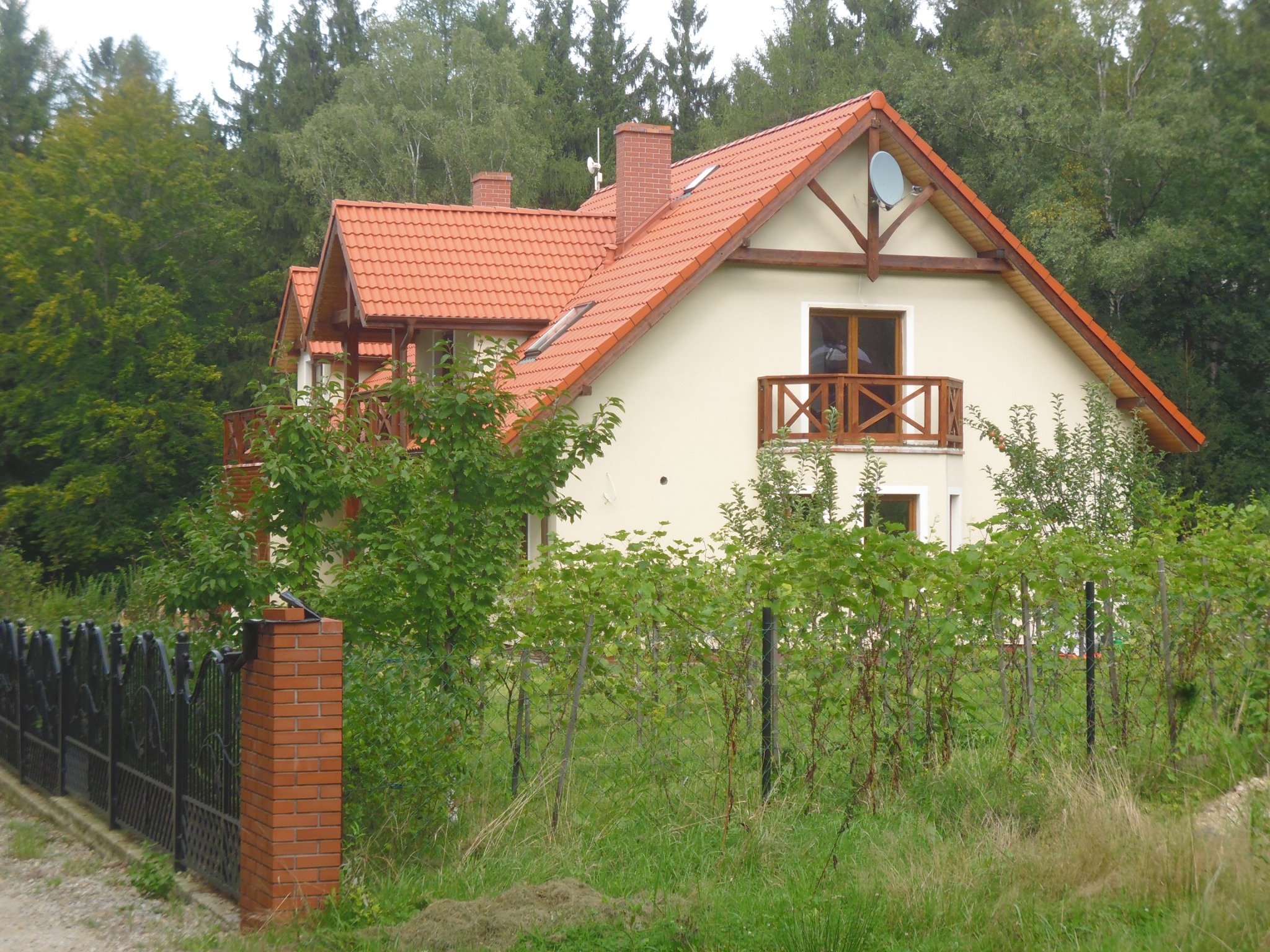 Dwupiętrowy dom z beżową elewacją, czerwoną dachówką, balkonem z drewnianą balustradą i anteną satelitarną na szczycie, otoczony zielenią i ogrodzeniem z ceglanymi słupkami, w powiecie wrocławskim.