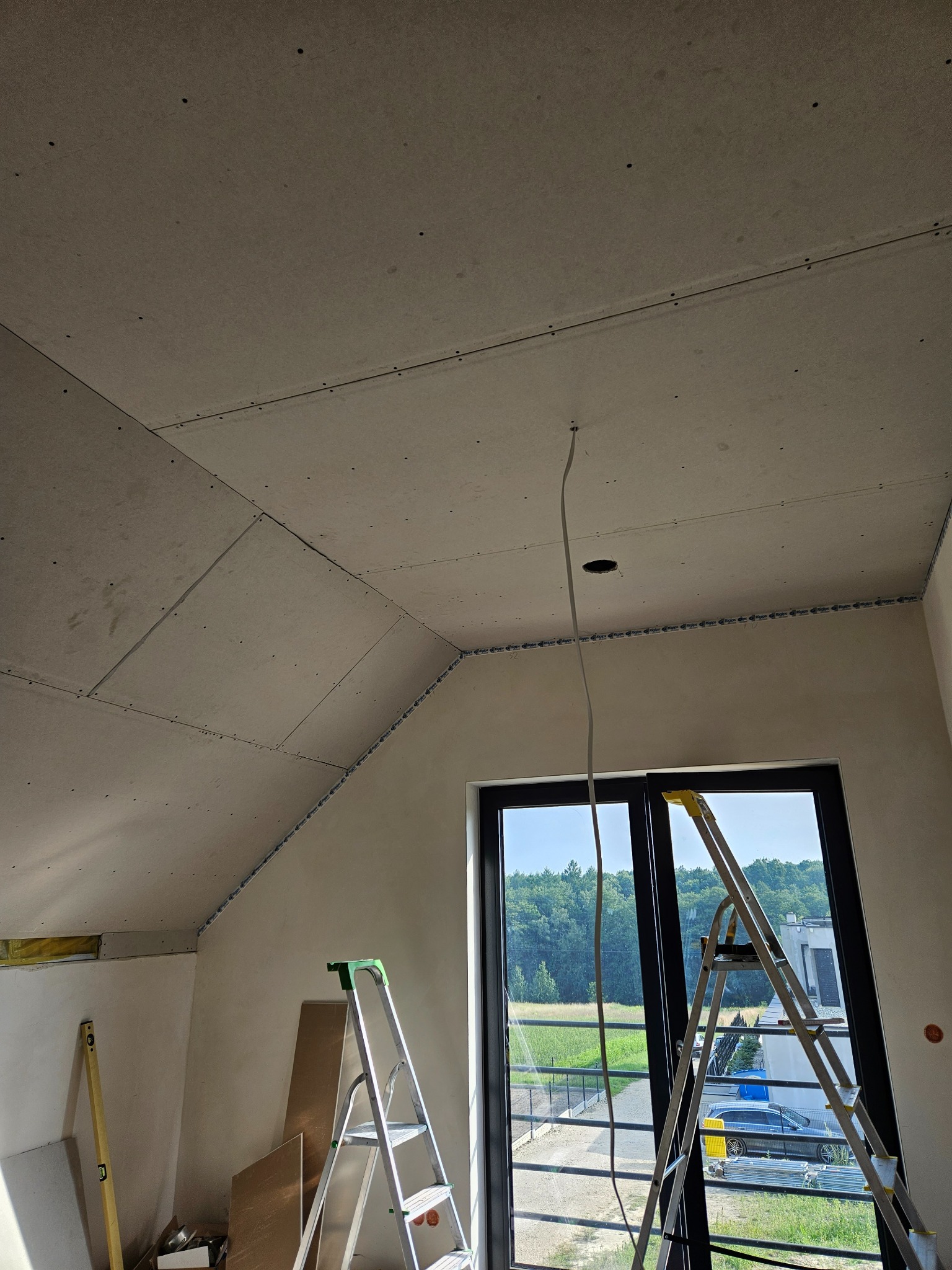 Poddasze w trakcie wykańczania: widoczne płyty na skosach, dwie drabiny opierające się o okno, wystający kabel z sufitu, poziomica oparta o ścianę.