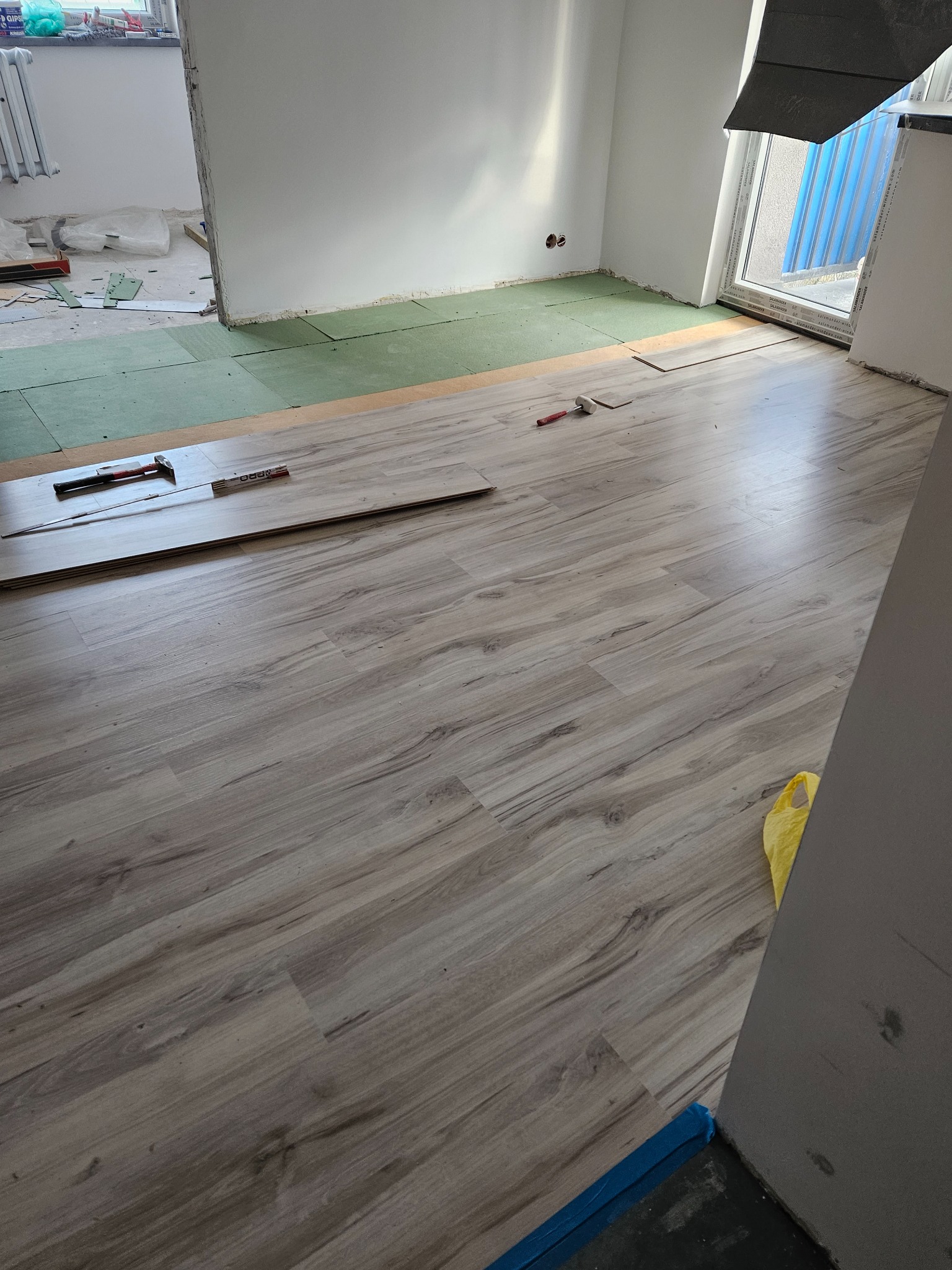 Układanie paneli podłogowych z widocznym podkładem, młotkiem gumowym i miarką w pomieszczeniu w trakcie remontu.