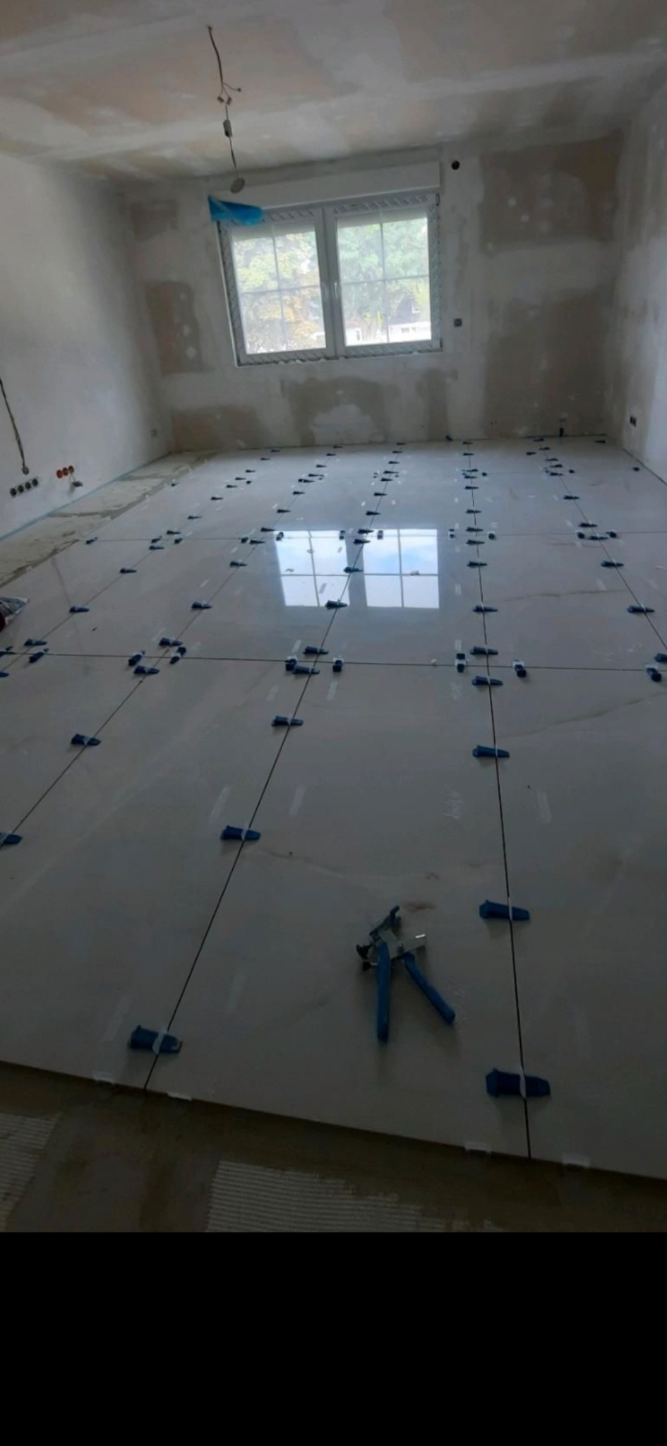 Wnętrze w trakcie układania jasnych, błyszczących płytek podłogowych z wykorzystaniem systemu poziomowania płytek; widoczne szczypce do klinów.